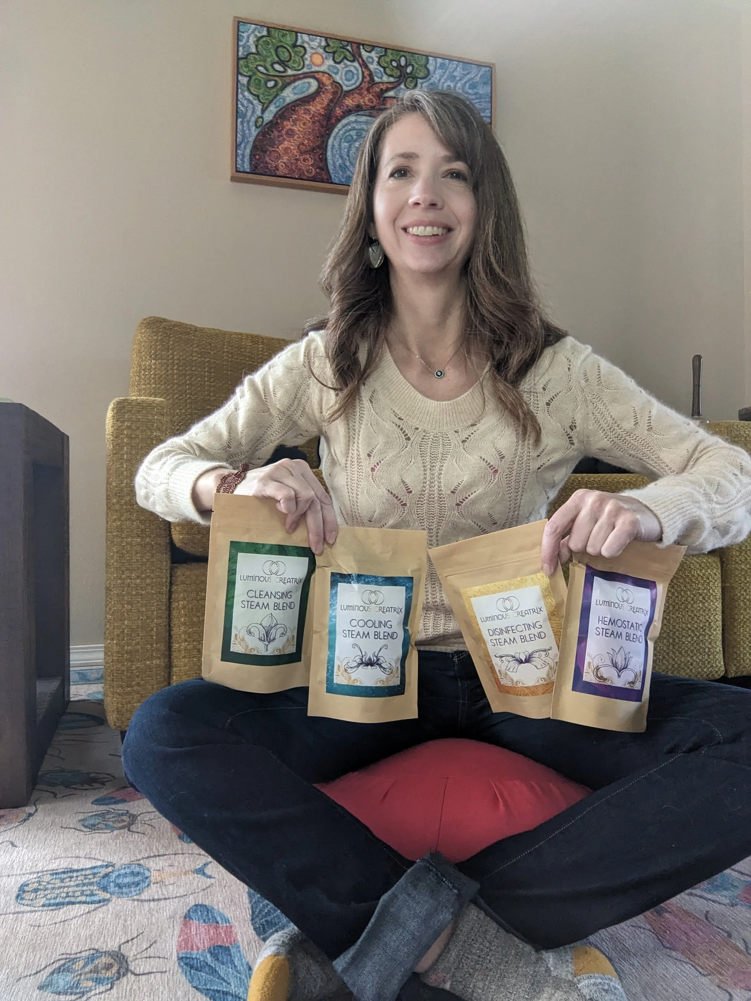 A woman sitting cross-legged on a rug with a red cushion in front of her. She is smiling and holding four packages of Luminous Creatrix steam blends labeled cleansing, cooling, disinfecting, and hemostatic. She is wearing a beige sweater, dark jeans, and yellow socks. Behind her is a yellow armchair and a colorful abstract tree painting on the wall.