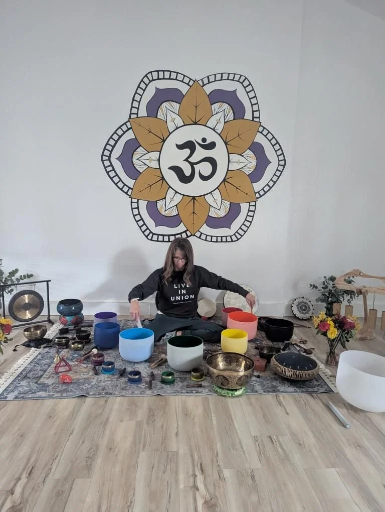 Woman sitting on a rug with singing bowls in front of her, arranging or playing them. Behind her is a large wall art with an 'Om' symbol surrounded by a decorative design featuring leaves and geometric shapes. There are more musical instruments and decorative items around her.