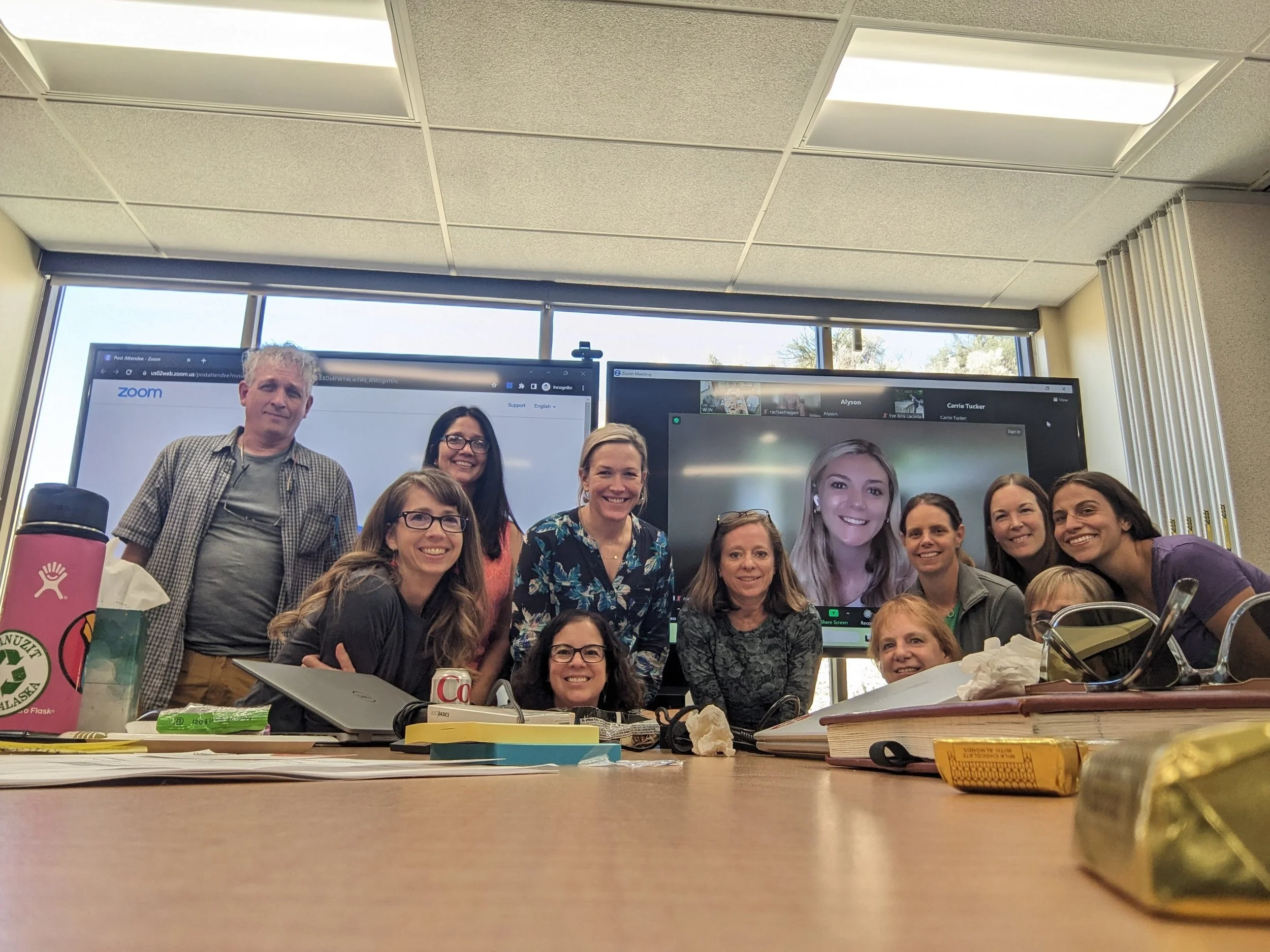 A group of people gathered in an office or conference room, smiling for a photo, with computer screens in the background, and cluttered desk with various items in the foreground.