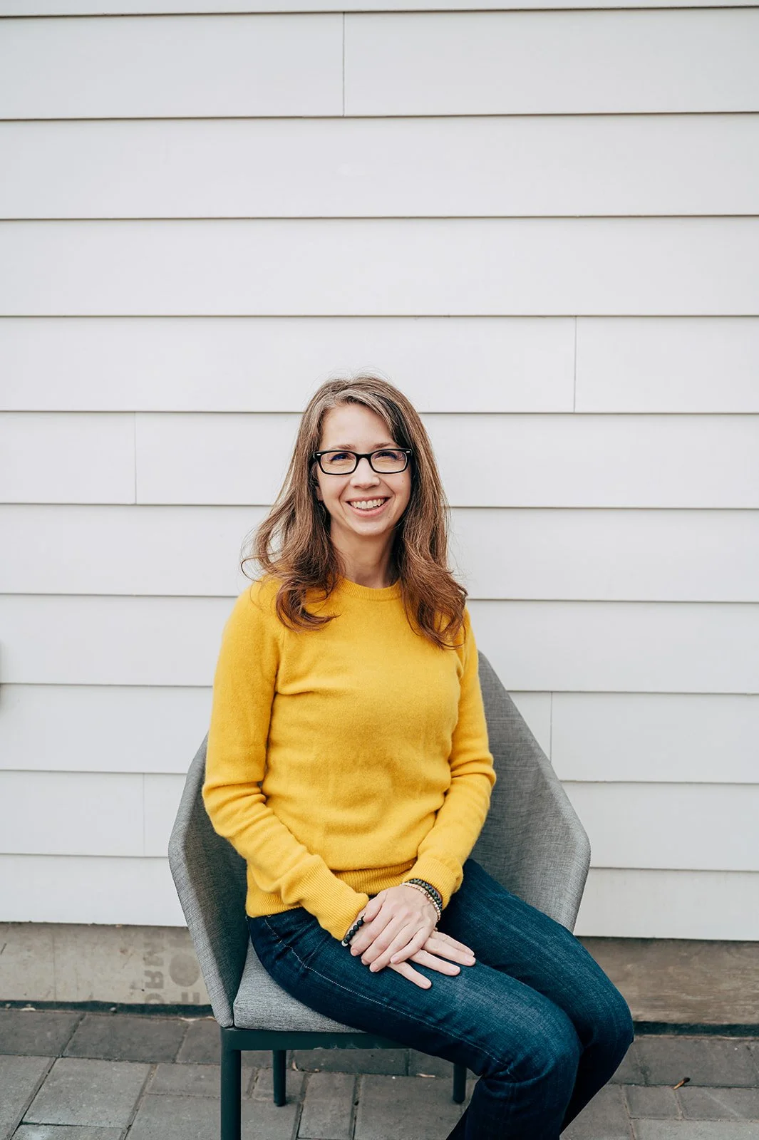 A woman with glasses and long brown hair sitting on a gray chair, smiling, wearing a yellow sweater and jeans, against a white horizontal panel wall.