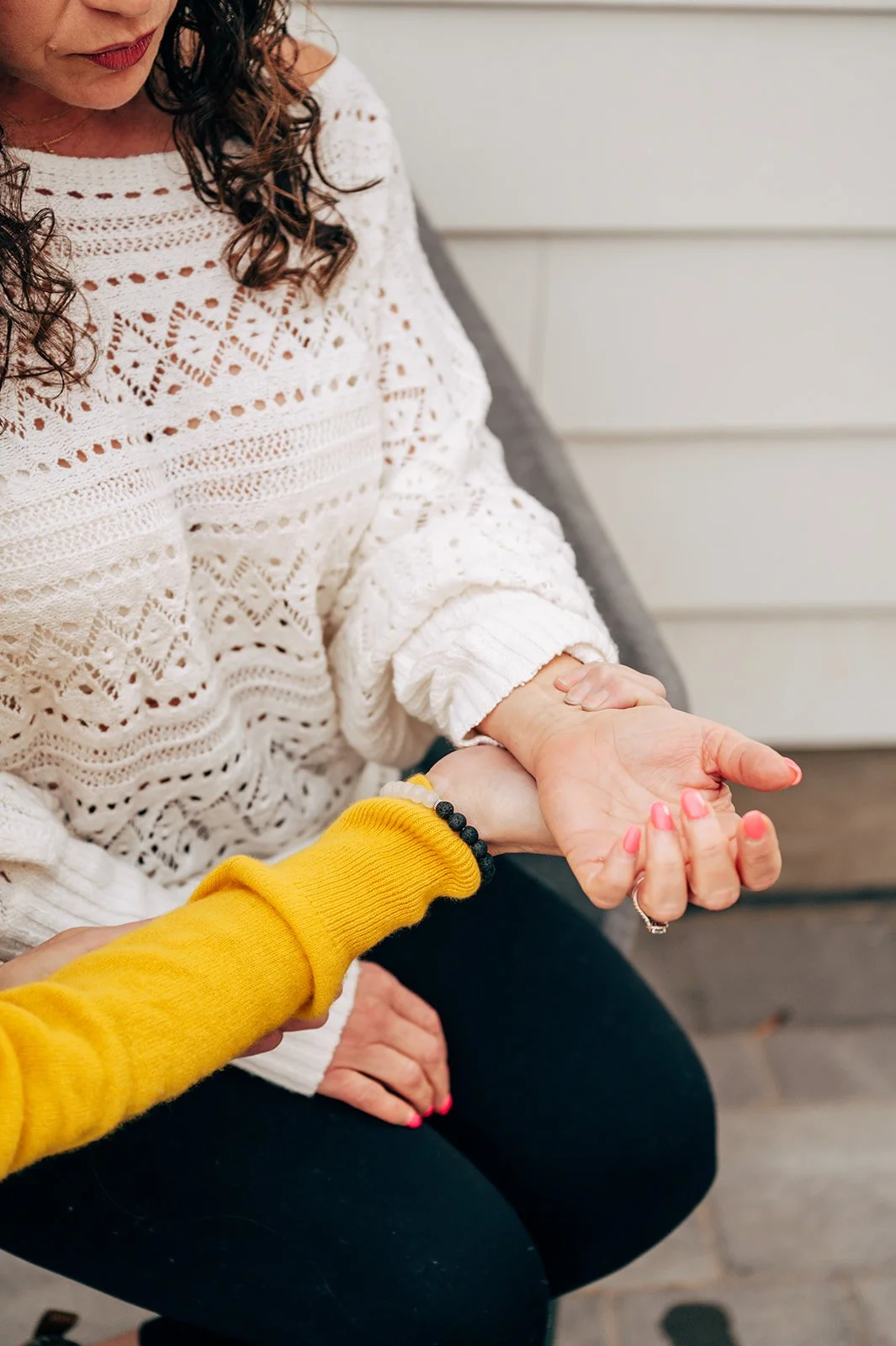 A woman with curly hair wearing a white crochet sweater, and another person in a yellow sweater holding her hand, with an outdoor setting in the background.