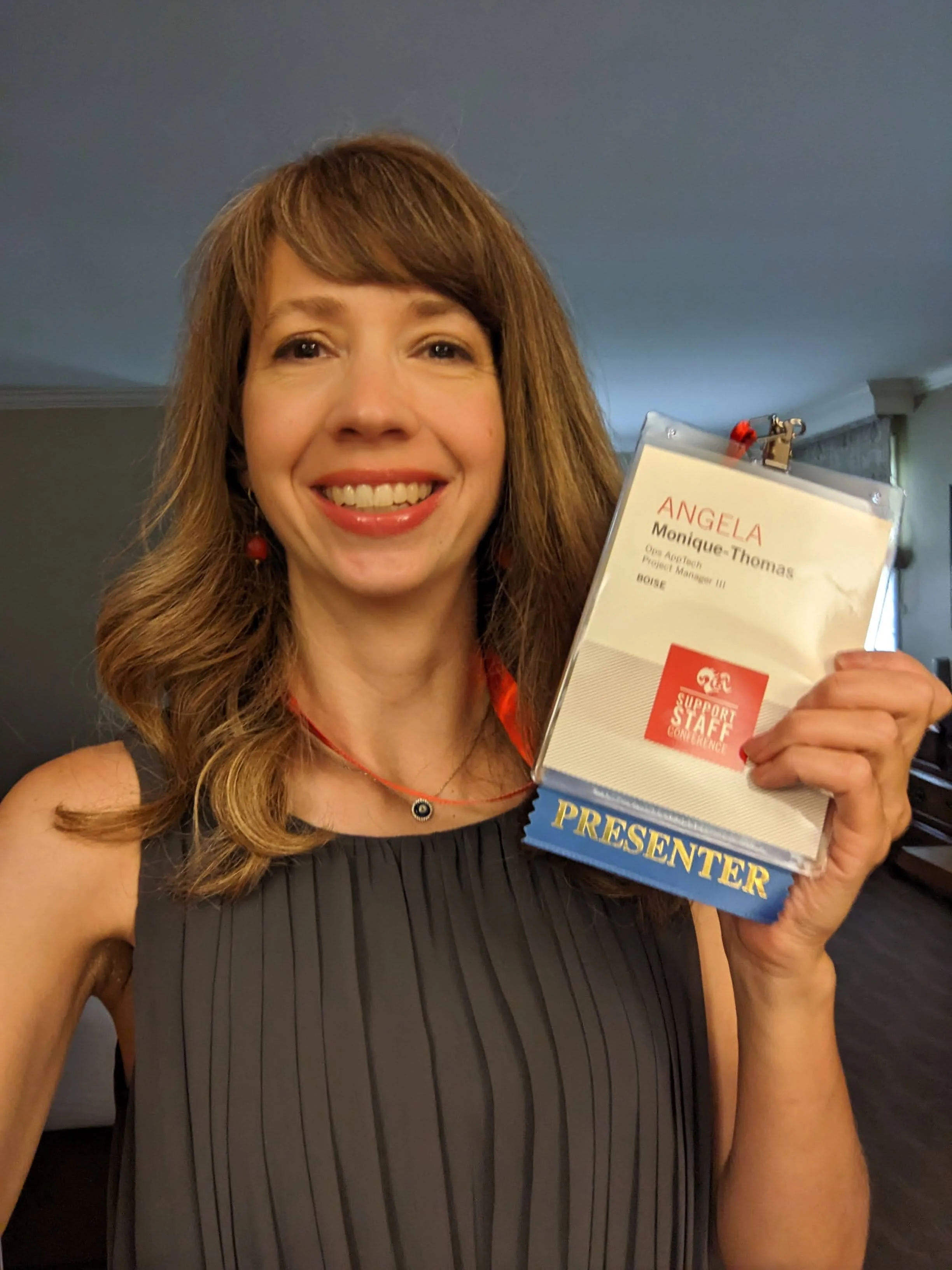 A woman with long reddish-brown hair smiling and holding a conference badge that says 'Angela Monique-Thomas, Open Project Manager III, Boise' and has a red sticker labeling her as a 'Support Staff Conference' presenter.