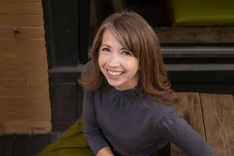 A woman with shoulder-length brown hair smiling at the camera, wearing a dark long-sleeve shirt, sitting in a cozy indoor space with wooden and brick accents.