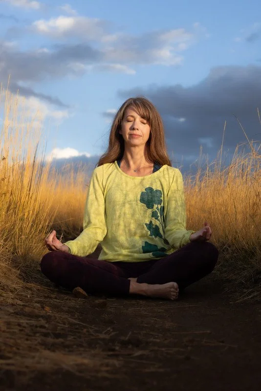 A woman practicing yoga outdoors in a field of tall grass during sunset, sitting cross-legged with hands resting on her knees in a meditative pose.