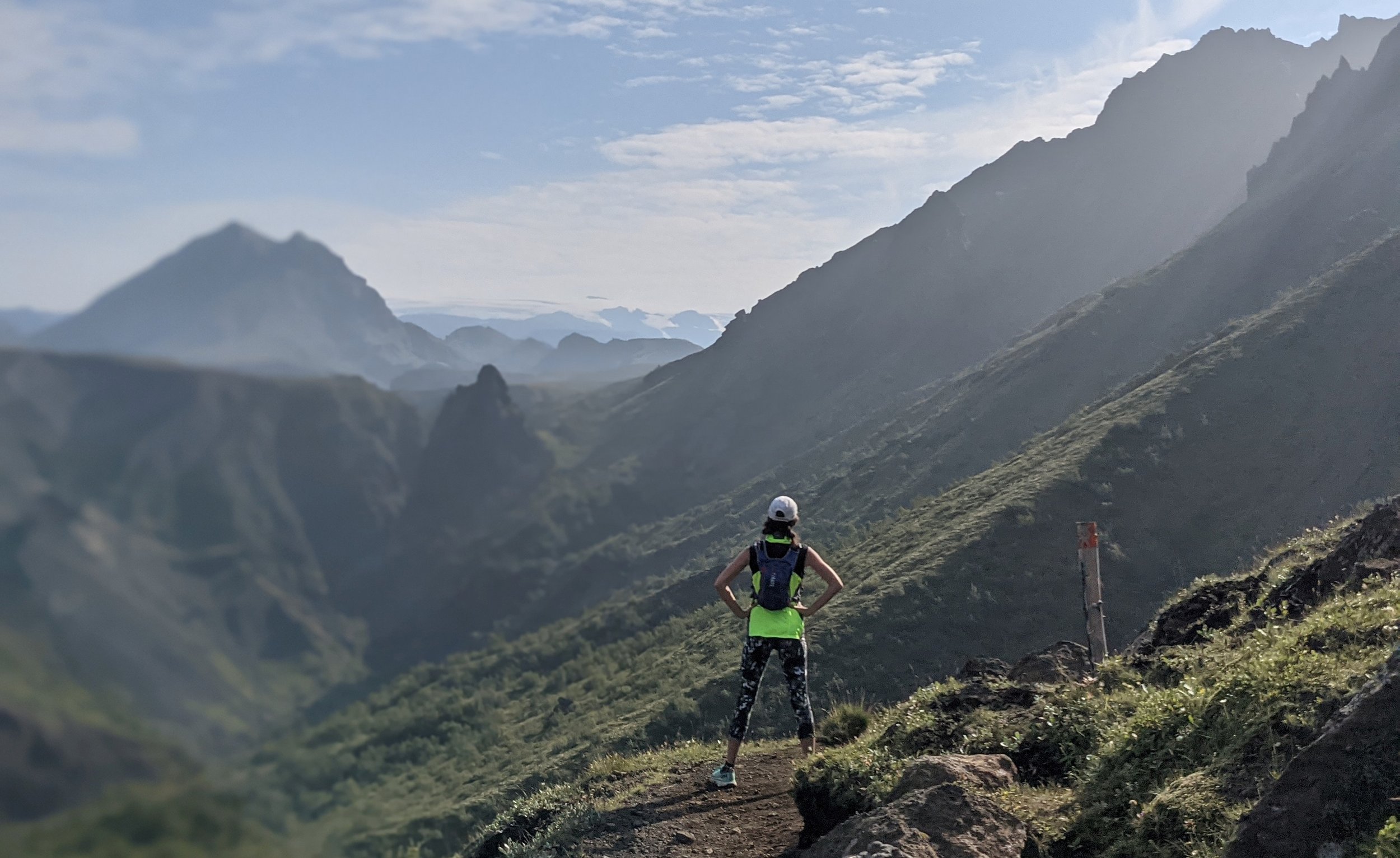 A person running on a mountain trail with a mountainous landscape in the background, wearing a white cap, a dark tank top, and a yellow-green skirt.