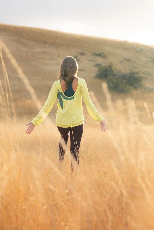 A young girl walking through a grassy field with open arms, wearing a yellow sweater and black leggings, during sunset.