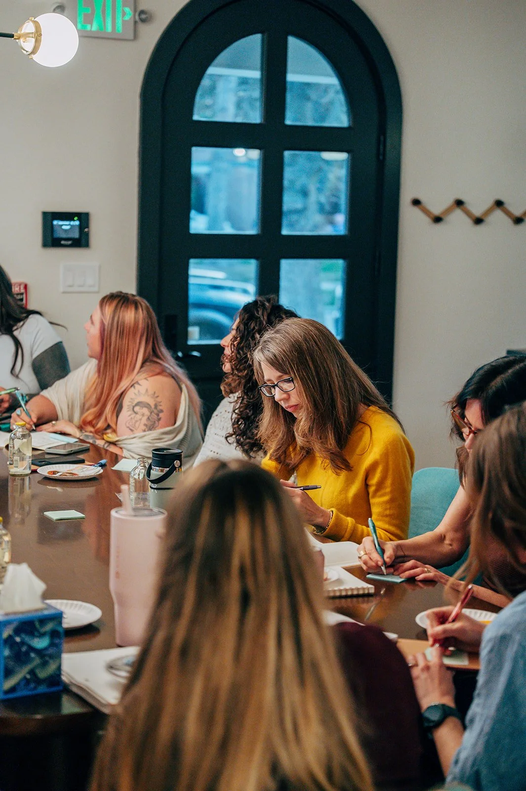 Women sitting around a wooden conference table, taking notes during a meeting in a room with black door and modern decor.