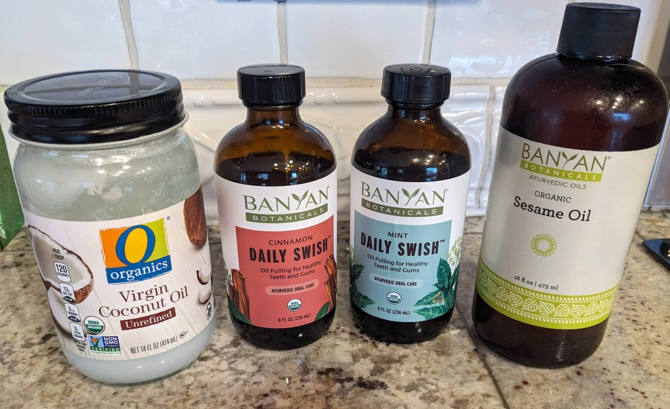 There are four bottles of oils on a kitchen counter, including coconut oil, cinnamon oil, mint oil, and sesame oil, with a tiled kitchen wall in the background.