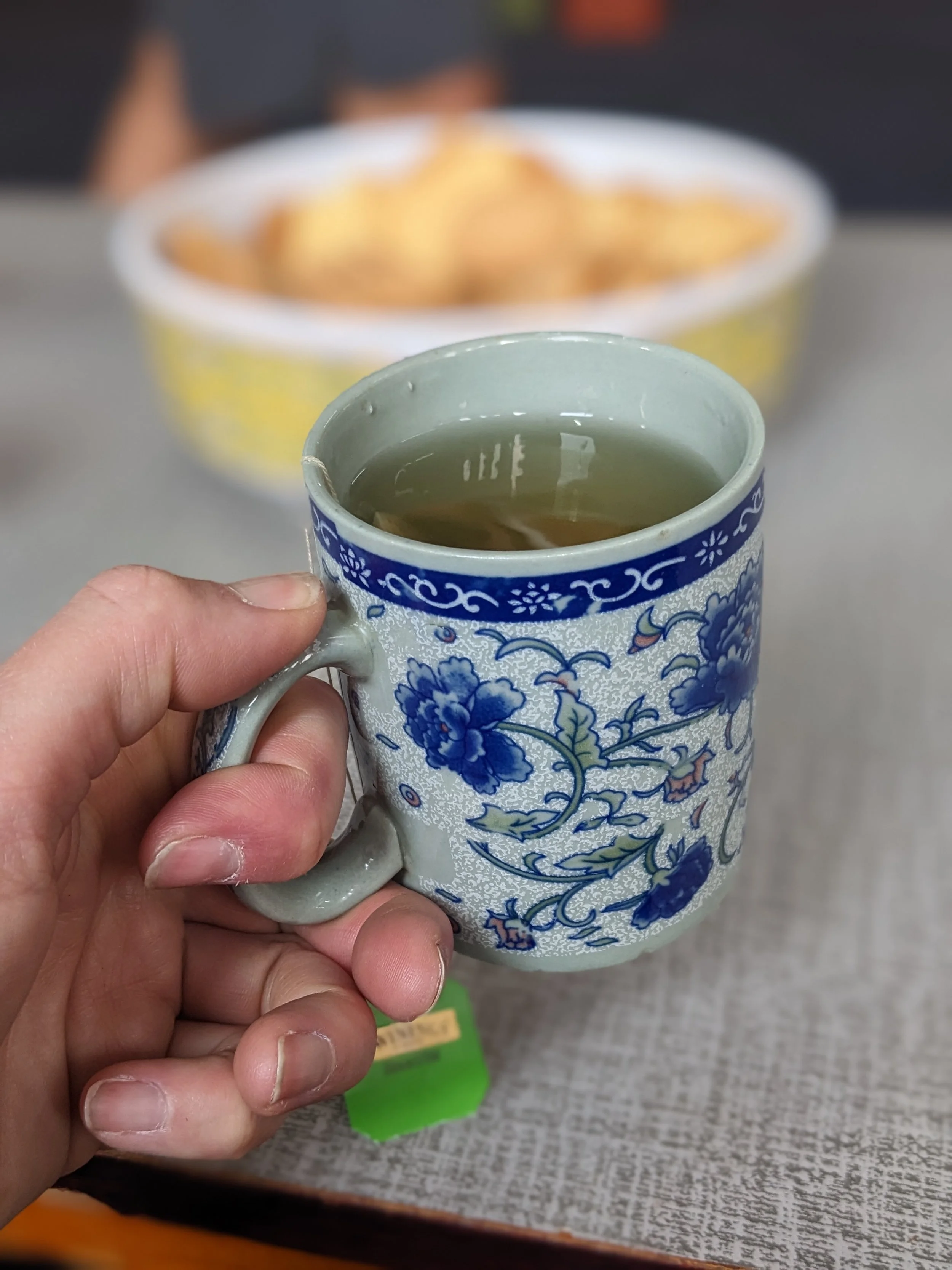 A person's hand holding a ceramic mug filled with a hot beverage, with a bowl of snacks in the background.