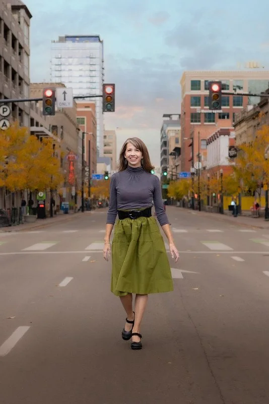 A woman walking down an empty city street with buildings and trees in the background, smiling and wearing a gray top and a green skirt.