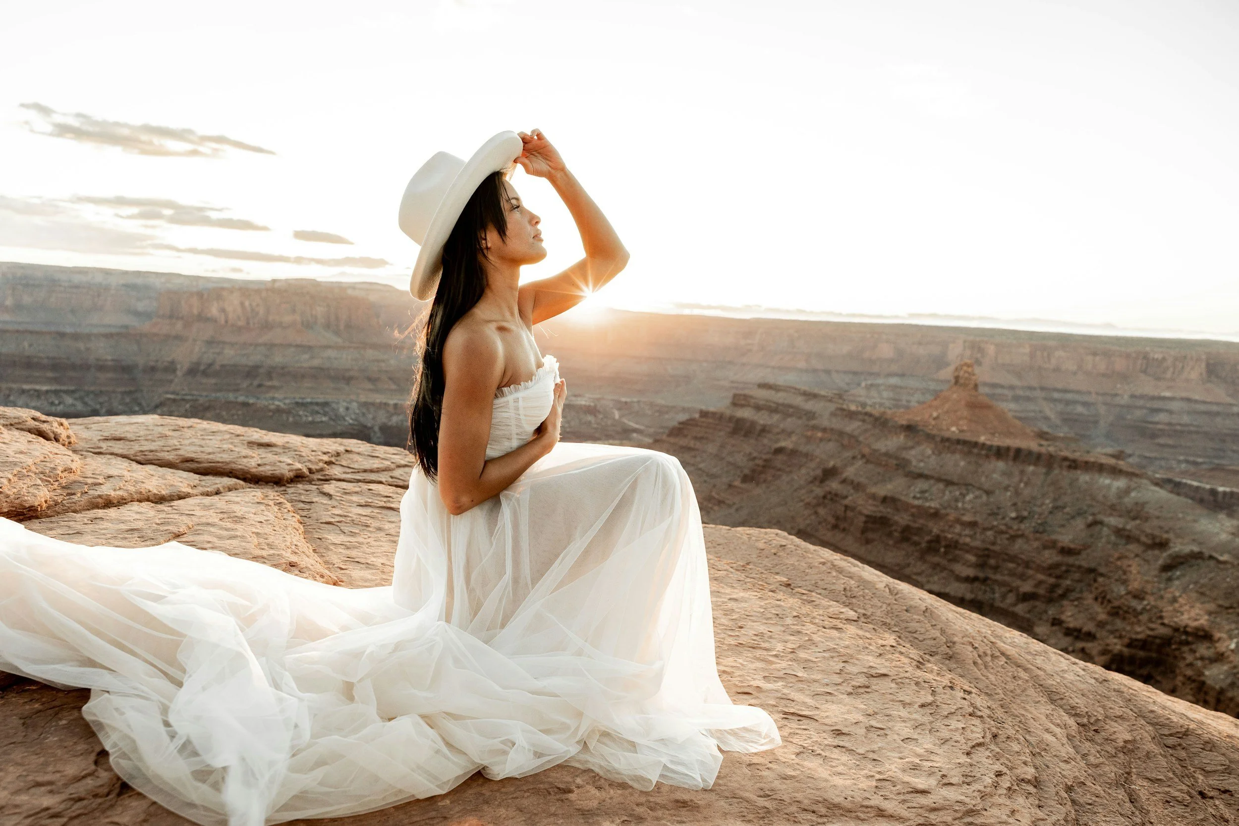 A woman in a white dress and wide-brimmed hat sitting on a rocks overlooking the canyon during sunrise or sunset.