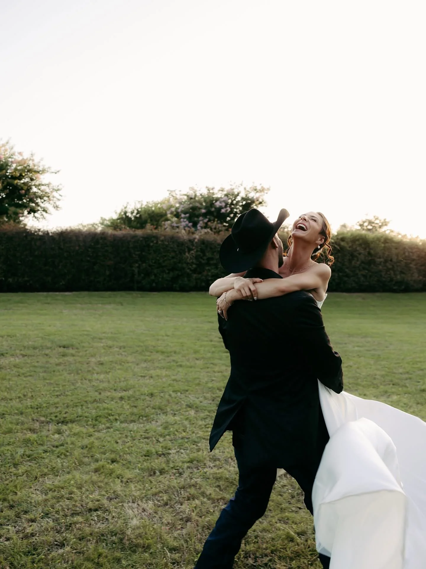 A bride and groom dancing in a grassy outdoor setting, with the groom wearing a black hat and suit, and the bride in a white wedding gown, both laughing and embracing.