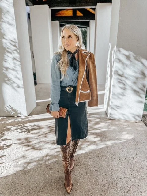 A woman with long blonde hair standing outdoors, smiling, wearing a denim shirt with a black necktie, a black leather skirt with a front slit, brown knee-high boots, and a brown and white jacket draped over her shoulder, holding a small clutch purse.