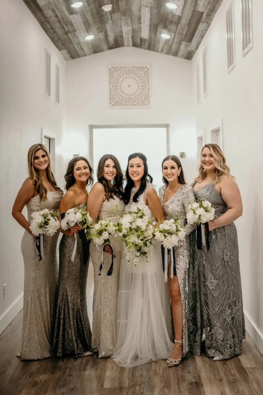 A bride and five bridesmaids standing in a hallway, all smiling and holding white bouquets, dressed in elegant dresses, with the bride in a white gown and the others in silver and gray gowns.