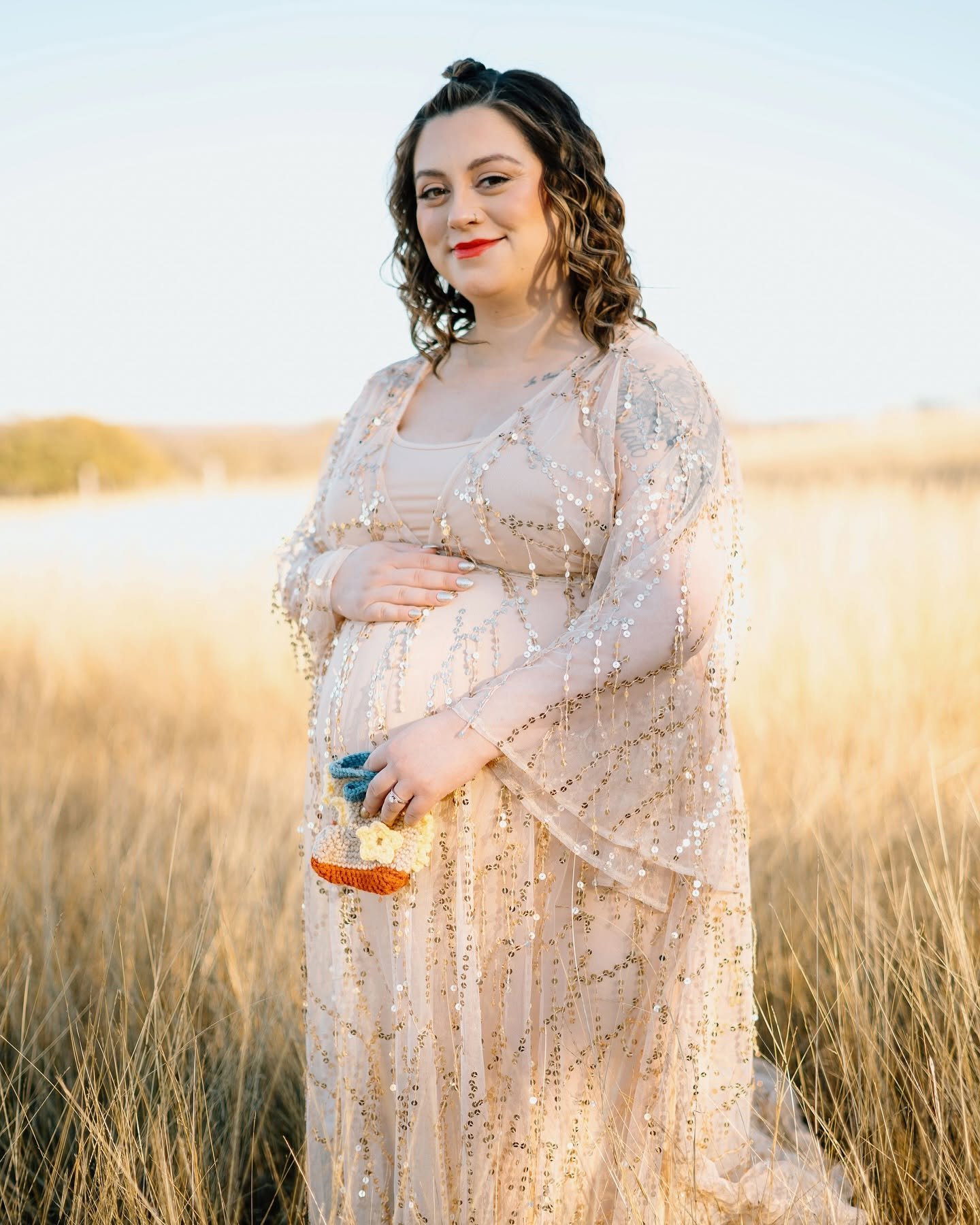 A pregnant woman with curly brown hair, wearing a sheer, beaded dress, stands in a golden field holding a crocheted item, smiling softly at the camera during golden hour.
