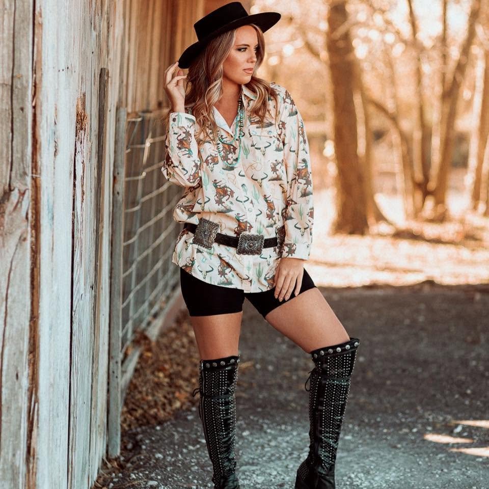 Woman wearing a cowboy hat, patterned shirt, black shorts, and knee-high studded boots, standing outdoors near a wooden fence with trees in the background.