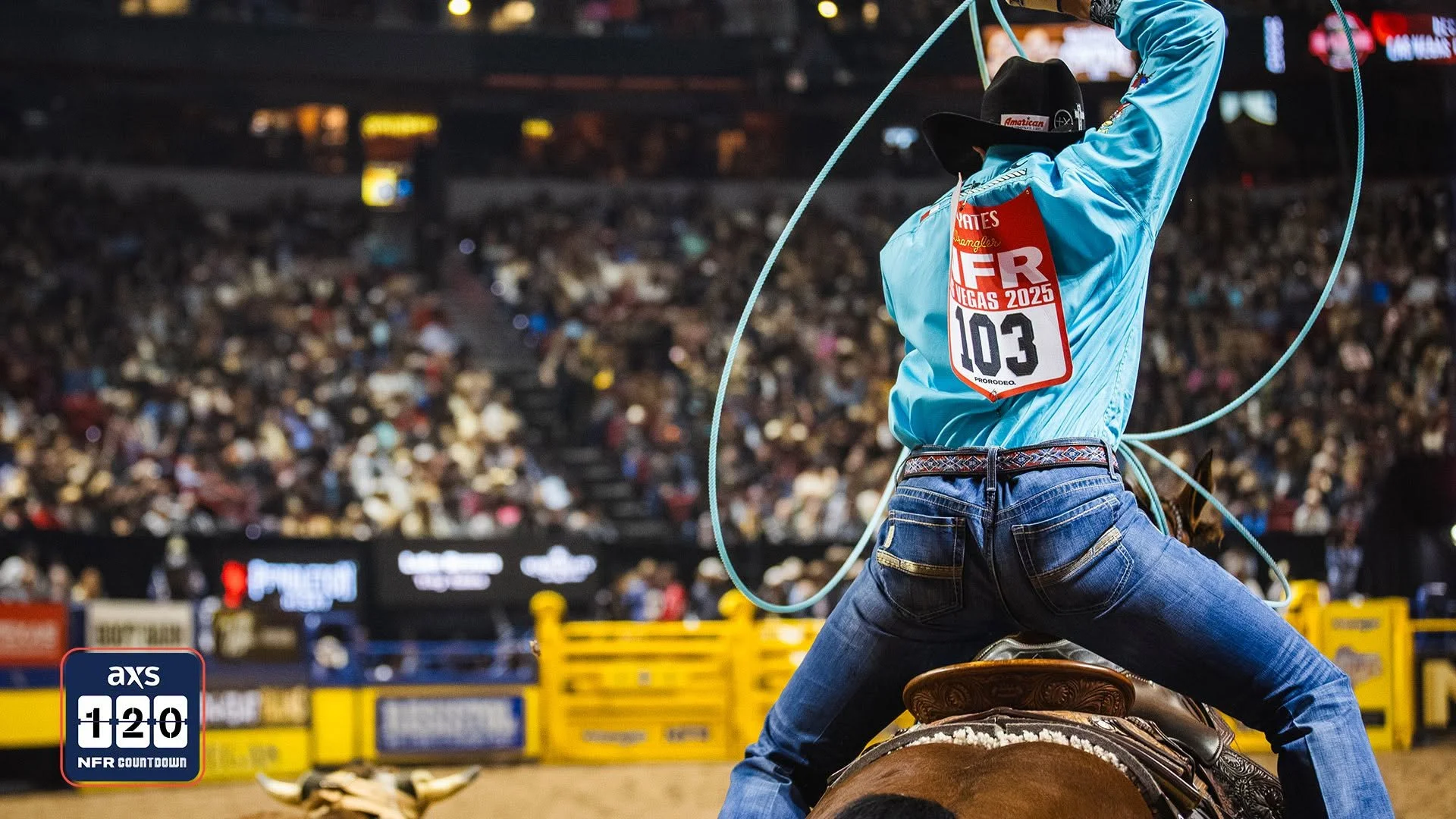 A rodeo cowboy wearing a black hat, light blue shirt, and jeans, holding a lasso, riding a bucking horse during a rodeo event, with a large crowd in the background.