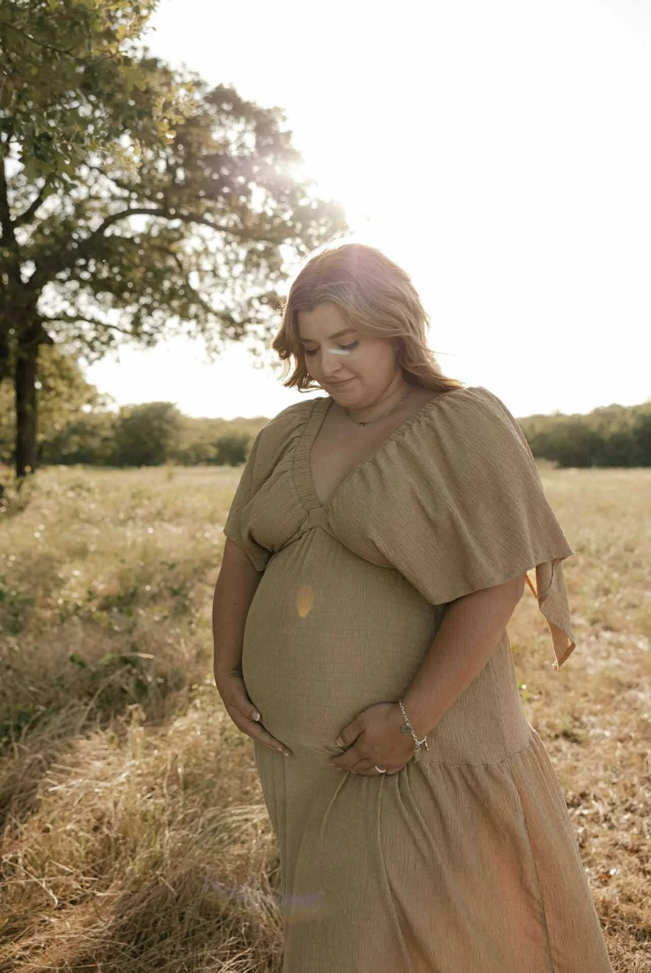 A pregnant woman with light brown hair in a field, holding her belly and looking down, dressed in a beige dress with short sleeves, with the sun shining behind her and trees in the background.