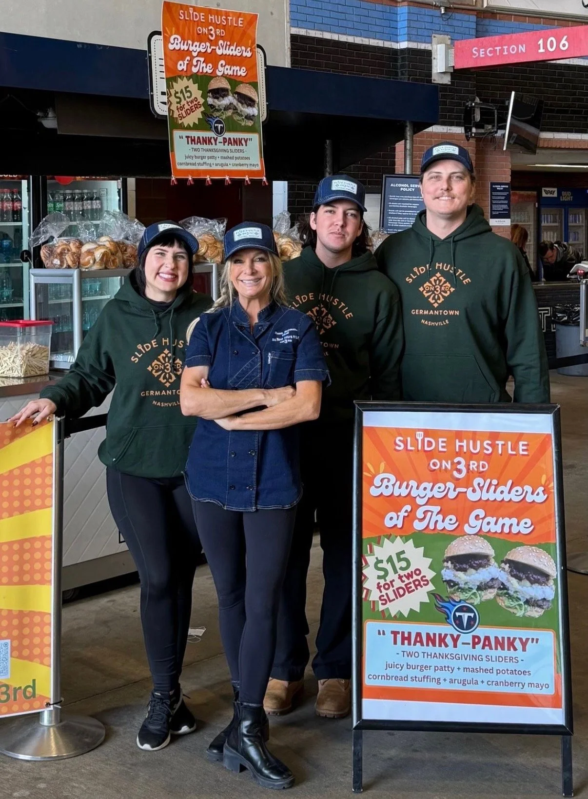Group of four people standing behind a signboard advertising burger sliders. Two women and two men are smiling and wearing green hoodies and blue caps. The signboard displays burger sliders with a promotion for two sliders and mentions ingredients like a juicy burger patty, mashed potatoes, and cranberry mayo. The background shows a brick building, and an orange banner promoting burger sliders