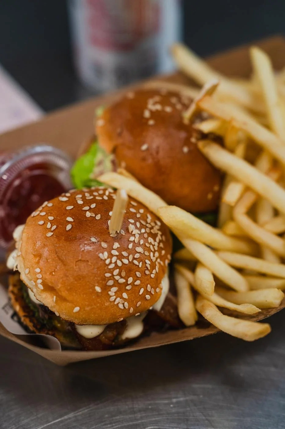 Two mini cheeseburgers with sesame seed buns, French fries, and a container of ketchup on a tray.