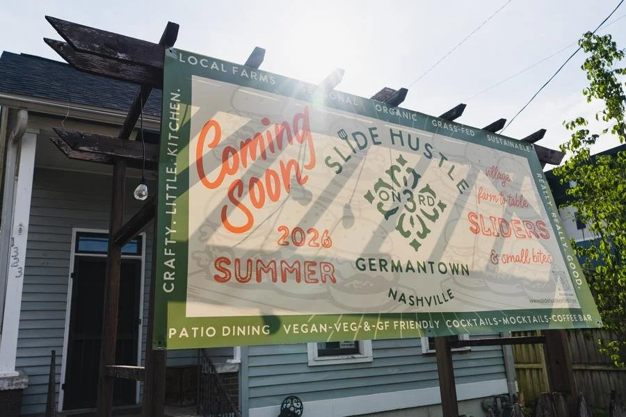 A large outdoor sign announcing the upcoming opening of a new restaurant or cafe. The sign mentions local, organic, grass-fed, and sustainable ingredients with a focus on summer, vegan, vegetarian, gluten-free options, patio dining, cocktails, mocktails, and coffee. It is located in Germantown, Nashville, and is coming soon in 2026.