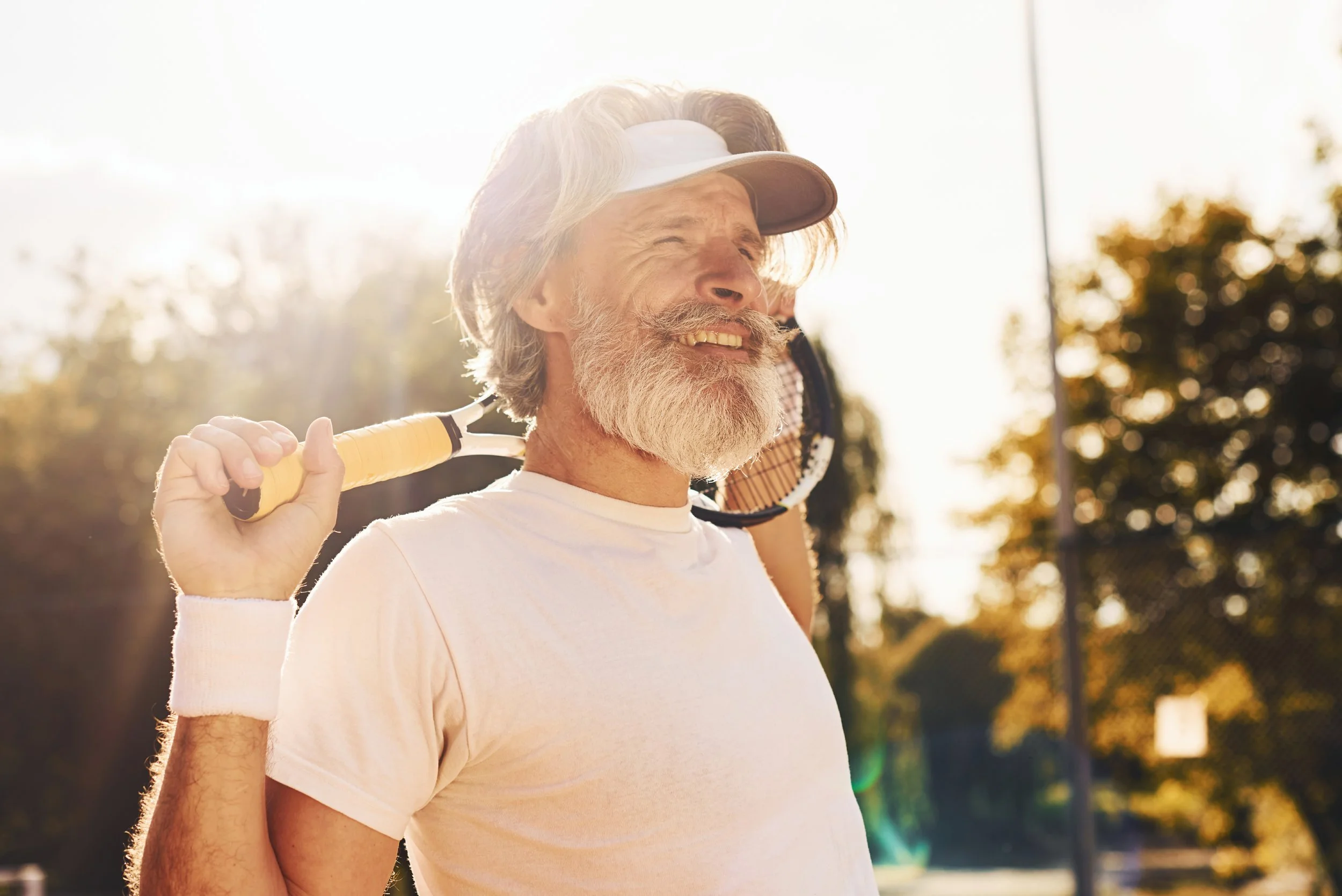 An older man with gray hair and a beard smiling while holding a tennis racket over his shoulder outdoors in sunny weather.