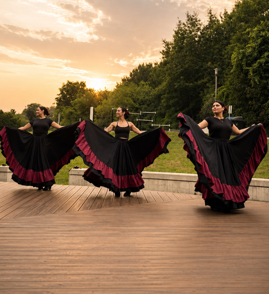 Three women in black dresses with red ruffles at the hem, dancing outdoors on a wooden stage at sunset, holding up their full skirts, with green trees in the background.