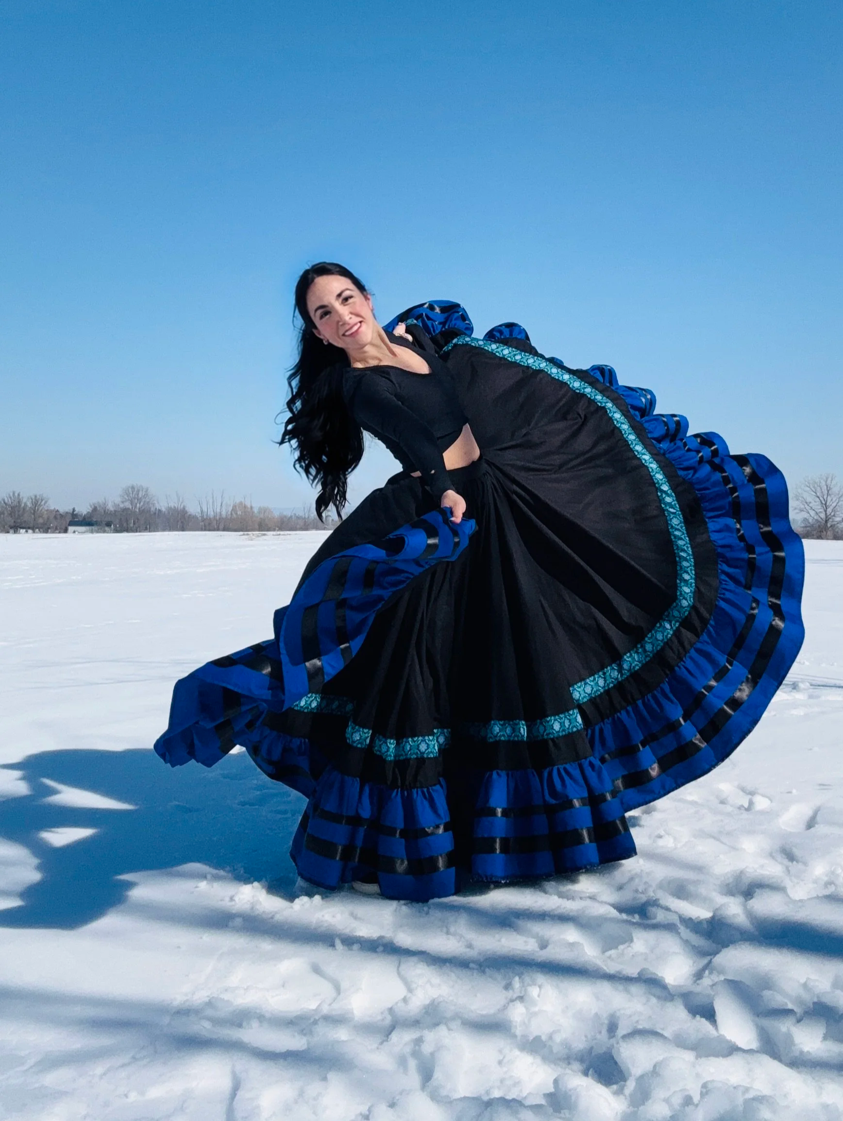 A woman laughing and spinning outdoors in a snowy landscape, wearing a black dress with blue and black striped ruffles around the hem.