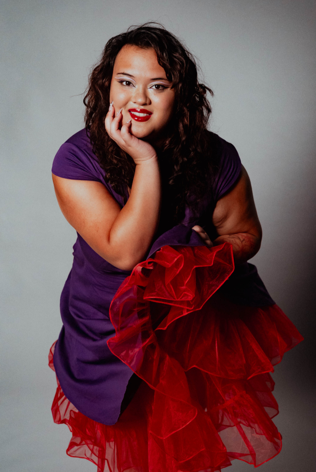 A young woman with curly dark hair, wearing a purple dress with ruffled red layers, posing with her hand on her face, smiling softly, against a plain gray background.