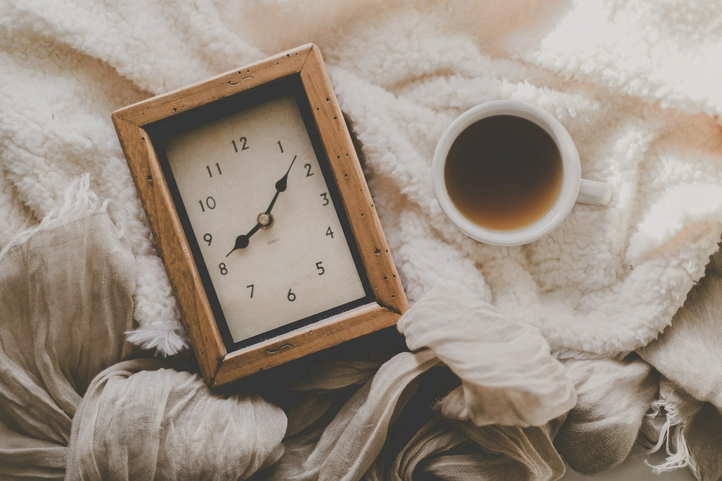 A person wearing beige pants and a beige sweater, sitting on a soft, fluffy white blanket. They are holding a small clock with a wooden frame displaying the time as 8:09 and a white mug filled with coffee or tea.