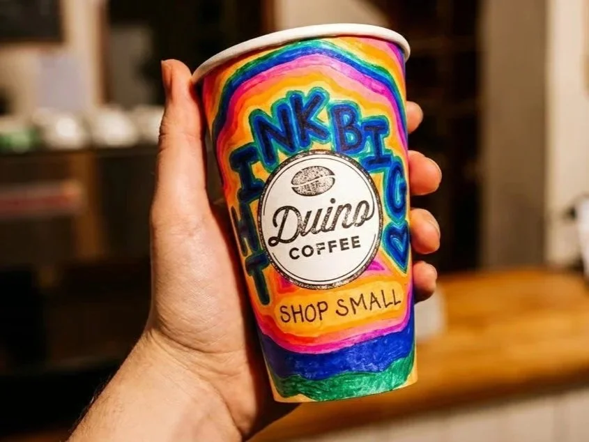 Colorful coffee cup with handwritten sign reading "Thankful, DUINO Coffee, Shop Small," held by a hand in a cozy indoor setting.