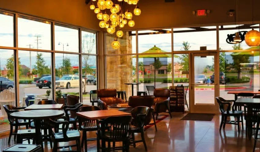 Interior of Duino Coffeehouse, wooden tables and chairs, hanging warm lights, patio umbrellas outside, and a large parking lot visible through the windows.