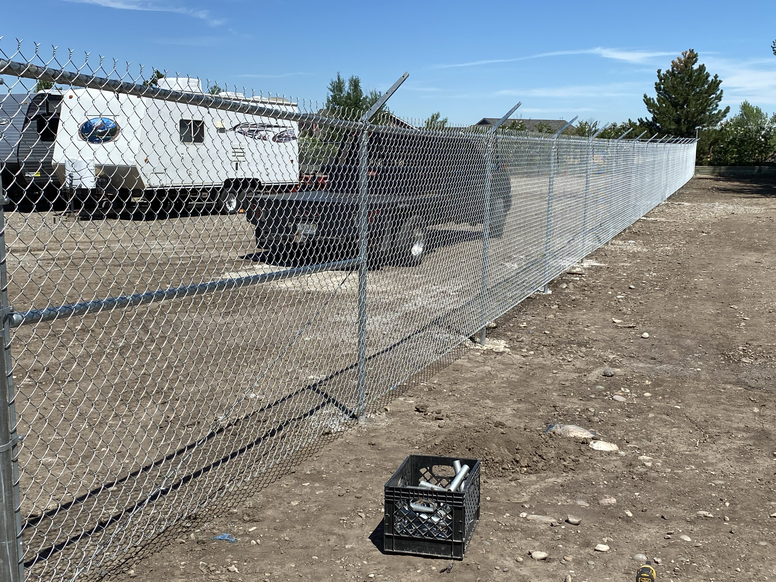 Long chain-link fence on a dirt lot with trees and houses in the background. A black crate with metal tools is on the ground near the fence. Clear blue sky overhead.