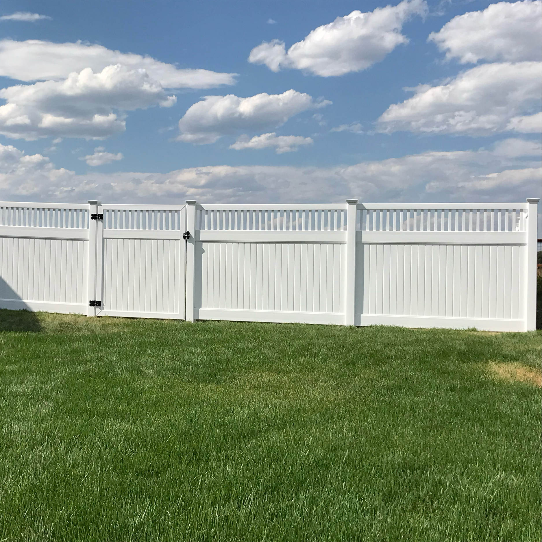 White vinyl fence with grass in front and blue sky with white clouds above.