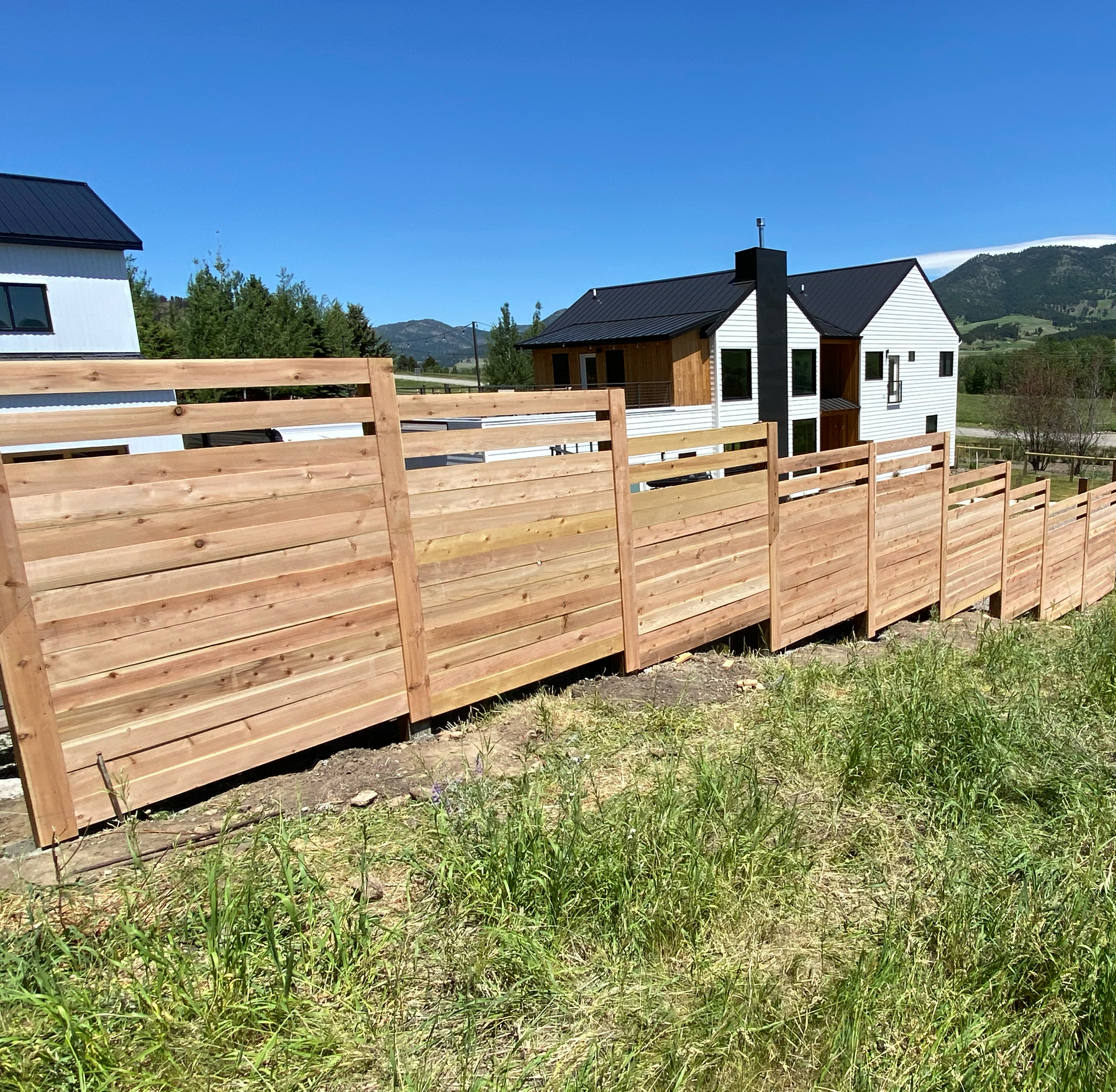 Newly constructed houses with black metal roofs and white siding, surrounded by a wooden fence and green hills in the background, on a clear sunny day.