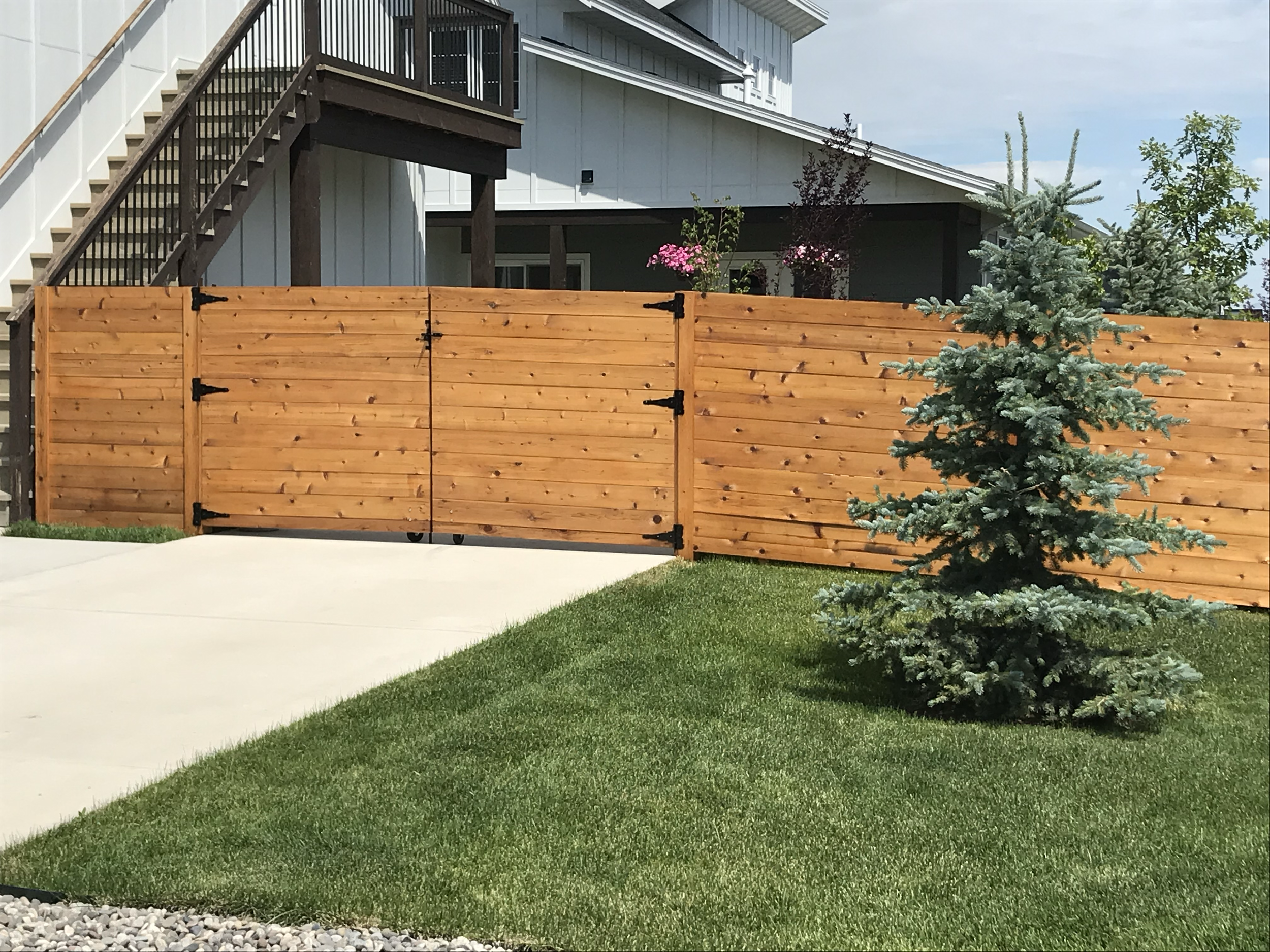 A backyard with a wooden privacy fence, green lawn, small decorative rocks, and a tall evergreen tree. In the background, there are side stairs of a house with a deck and plants with pink flowers.