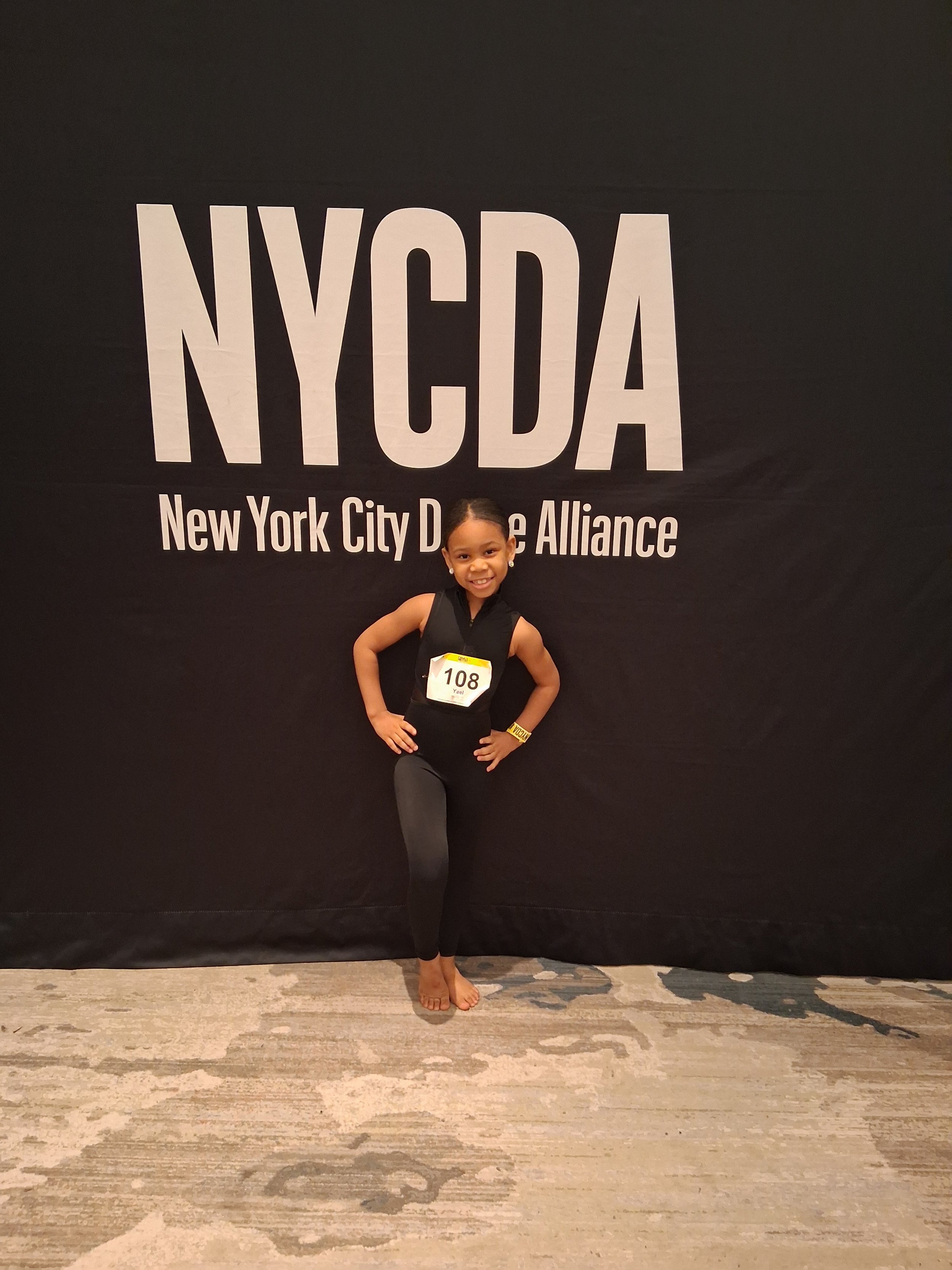 A young girl dressed in black fitness attire poses with her hands on her hips in front of a backdrop with the text 'NYCDA' and 'New York City Dance Alliance'. She is smiling and wearing a competition number '108' on her chest, standing on a light-colored rug.