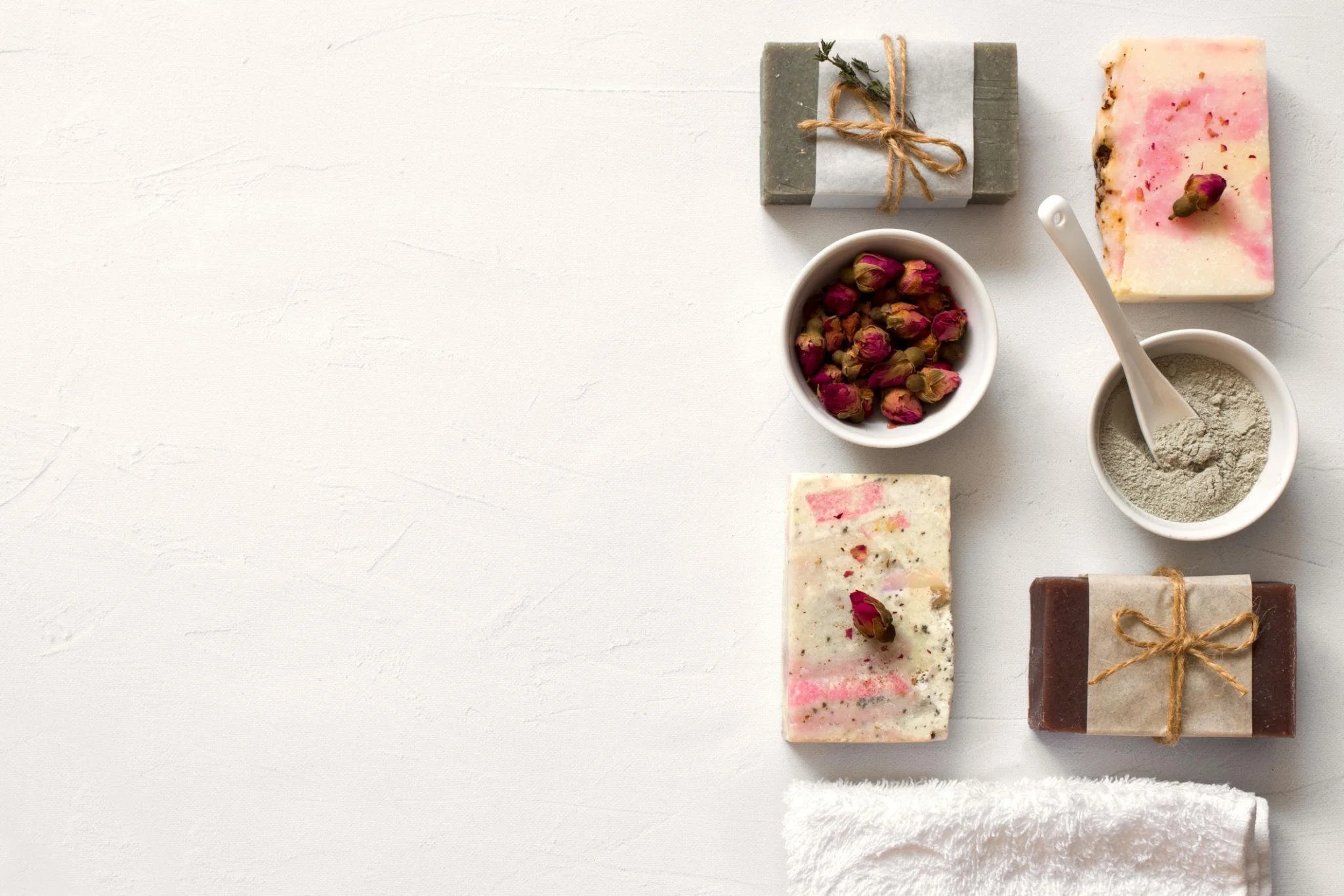 A flat lay of colorful handmade soap bars, a small bowl of dried rosebuds, a small bowl of gray powder with a spoon, and a small gift wrapped in rustic paper and tied with twine, arranged on a white textured surface.