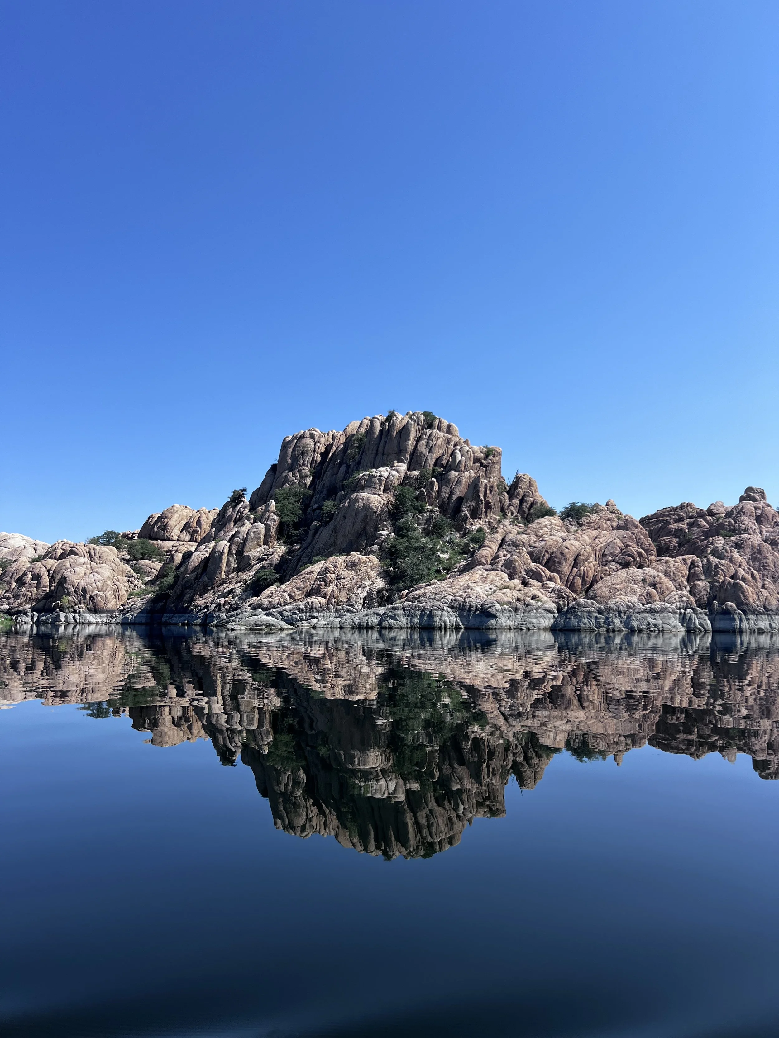A rocky mountain with trees and a clear blue sky reflected in calm water.