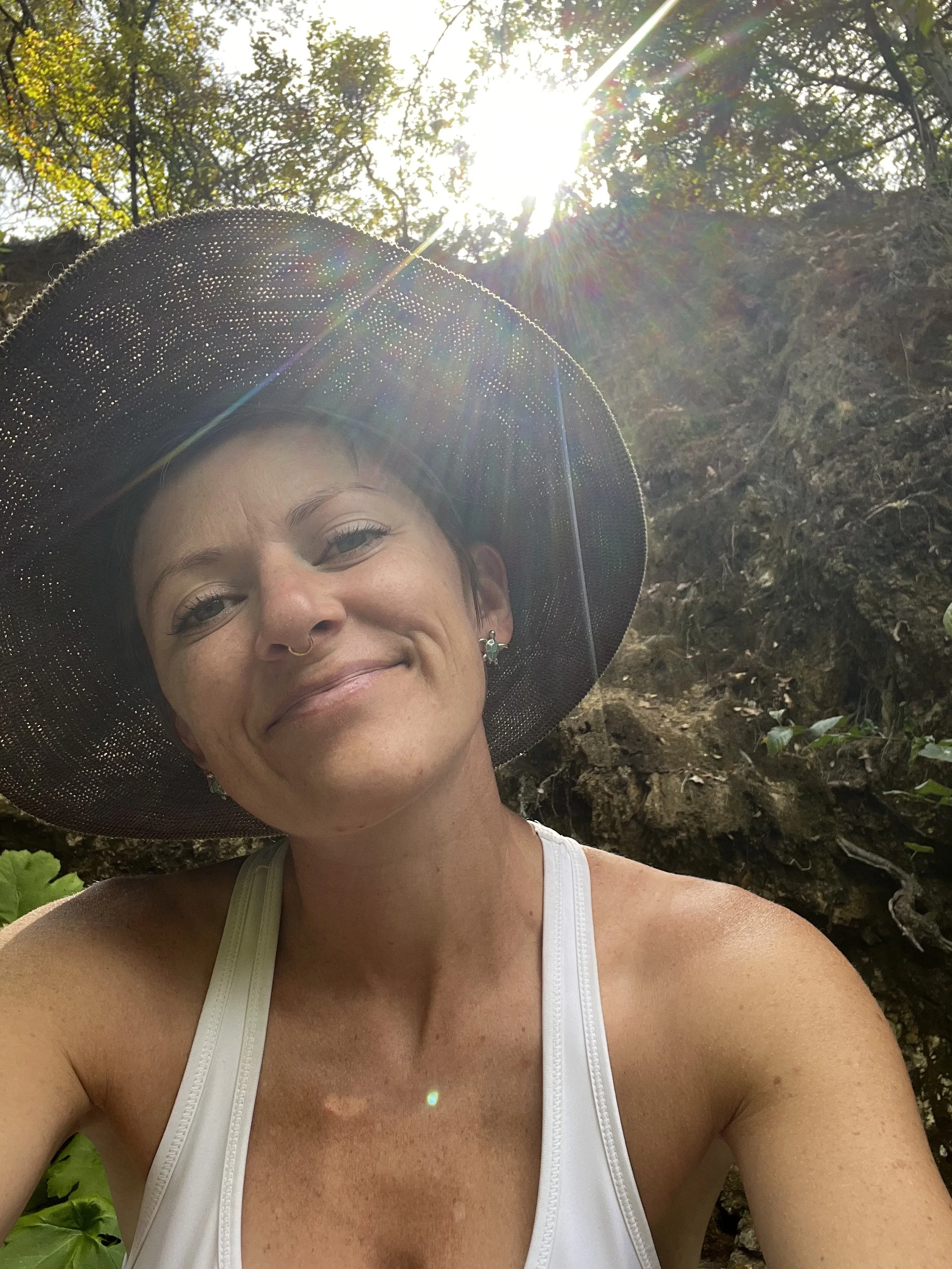 A woman smiling outdoors wearing a black wide-brimmed hat, white tank top, and earrings, with sunlight shining through trees in the background.