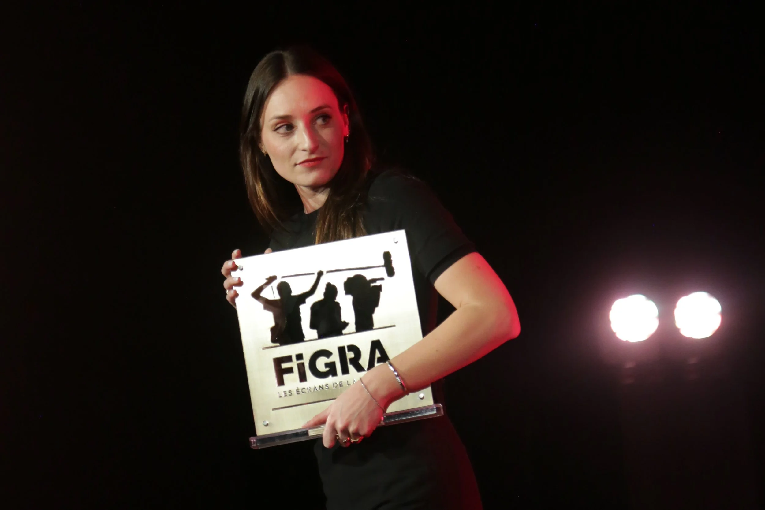 A woman with brown hair and a black shirt holding a sign with black silhouettes of people and text, against a black background with bright lights.