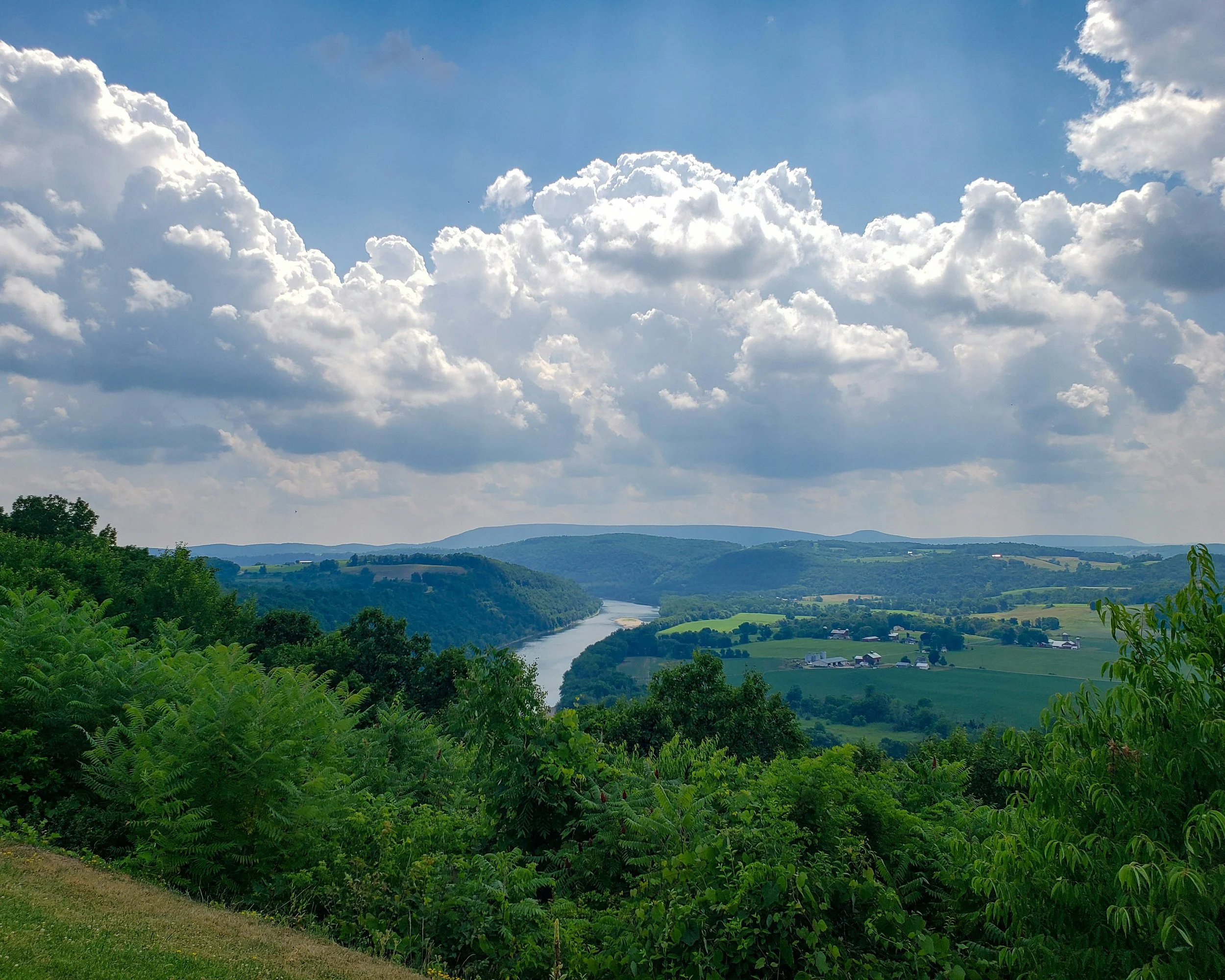 A scenic landscape of rolling green hills, a river winding through the valley, and a partly cloudy blue sky above.