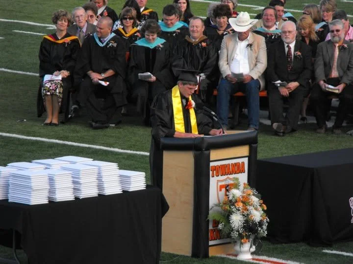 A graduation ceremony taking place outdoors on a football field, with a graduate speaking at a podium decorated with flowers, and an audience of faculty and guests seated behind him.