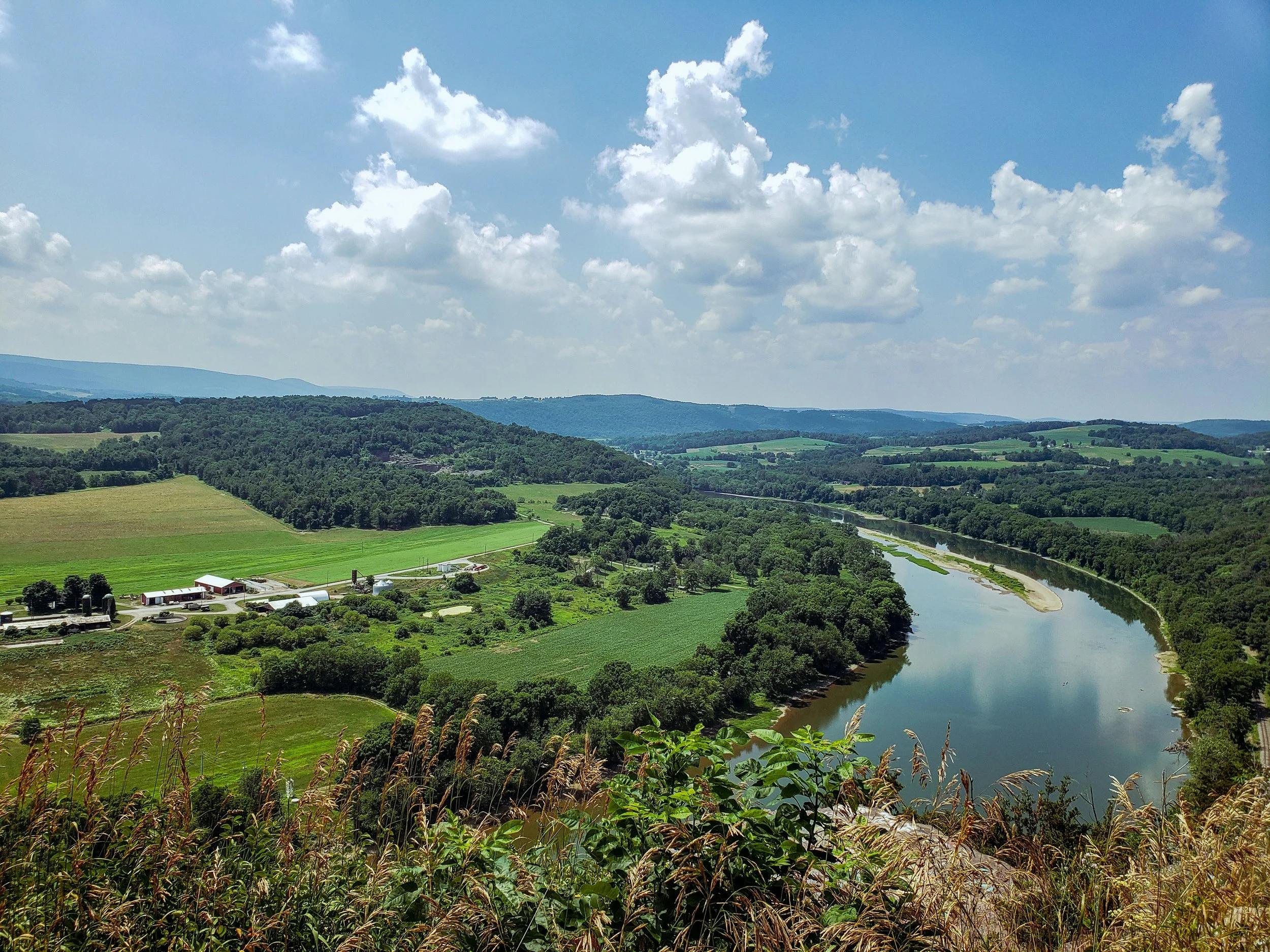 Scenic landscape of rolling green hills, a river winding through the countryside, and a partly cloudy blue sky.