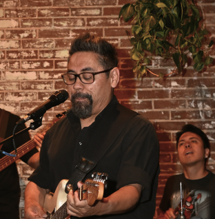 A man with glasses and a beard playing an acoustic guitar and singing into a microphone, with another person in the background, in a setting with a brick wall and green plant.