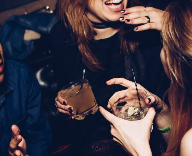 Two women holding glasses of mixed drinks, engaging in a conversation, smiling with one woman touching her lips with her finger.