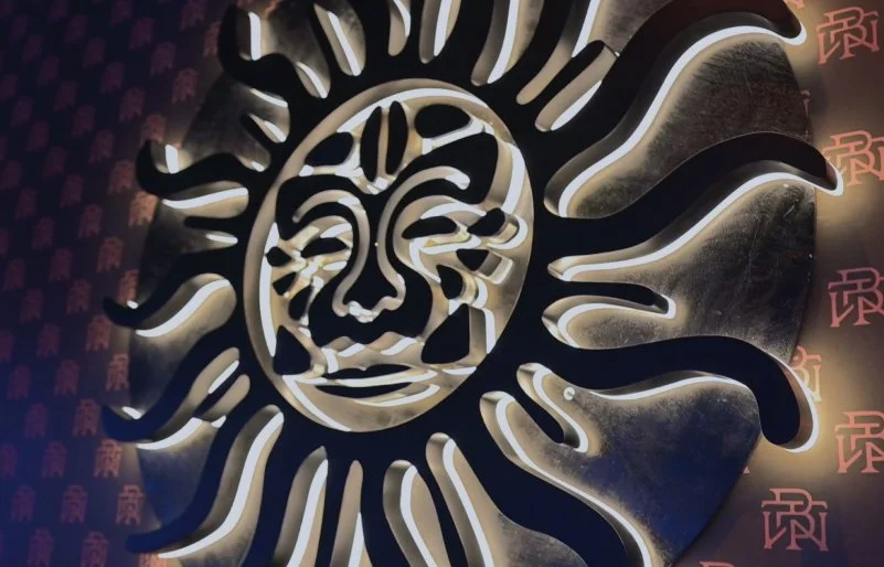 A carved and back-lit stylized sun design, representing the membership concept, on a textured wall at By The Wayside cocktail bar in Houston.