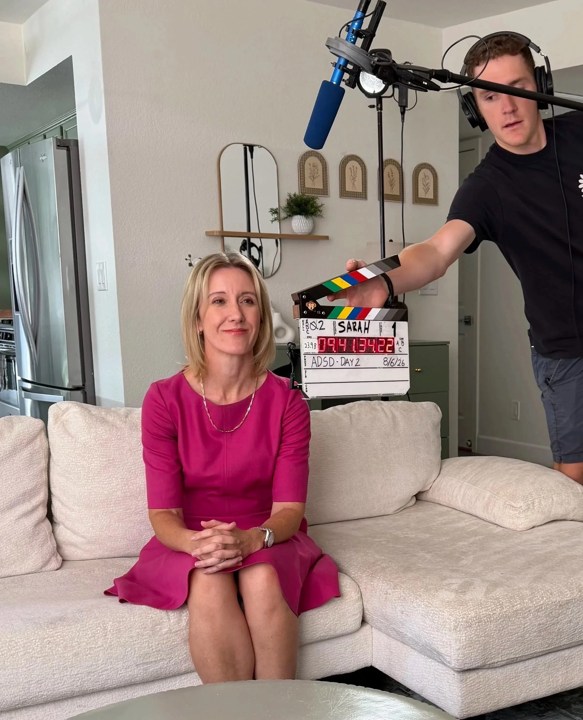 Sarah Peaceful sitting on a beige couch, wearing a pink dress, smiling. Behind her, a man is holding a clapperboard with the label 'SARAH', and a video recording device with a microphone is pointed at her.
