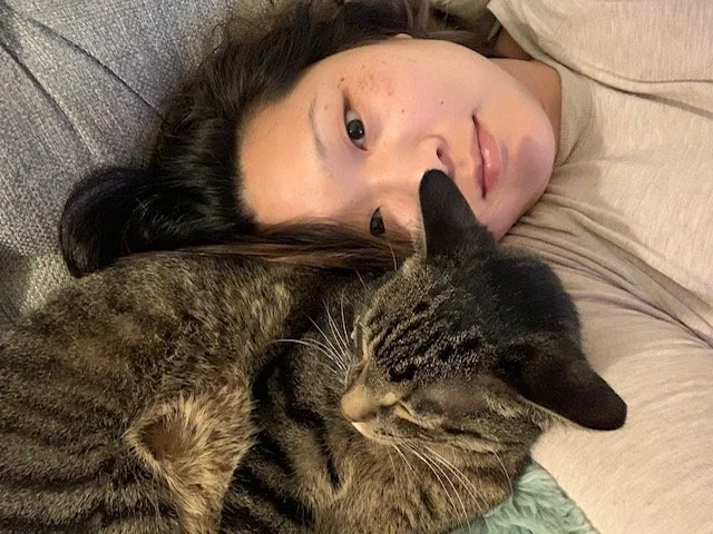 A Korean woman in her 30s lying on a couch with a brown tabby cat leaning on her face.