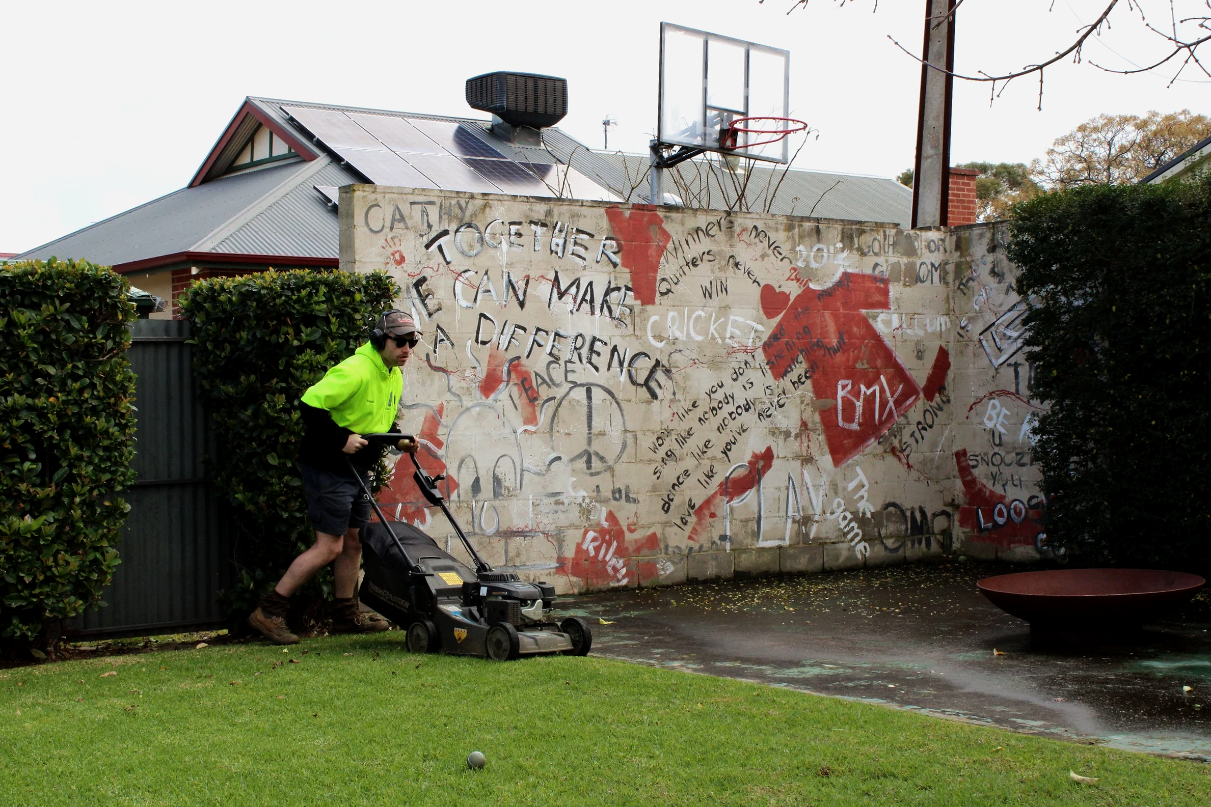 A man in a neon yellow jacket mowing the lawn near a graffiti-covered wall with a basketball hoop mounted on top, a house with solar panels, and trees in the background.